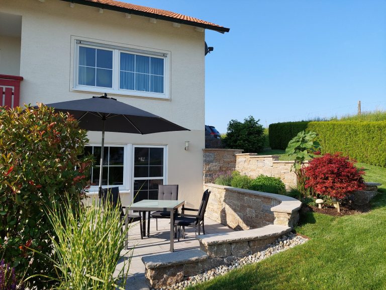Ferienwohnung Haisterkirch Ein sonniger Gartenbereich mit einer Terrasse, Tisch und Stühlen neben einem Haus.